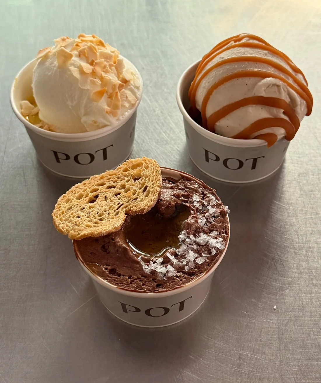 Tres vasos Pot sobre un mostrador de acero: helado de yogur con coco tostado, helado de vainilla con caramelo salado y mousse de chocolate con aceite, sal y una tostada de pan